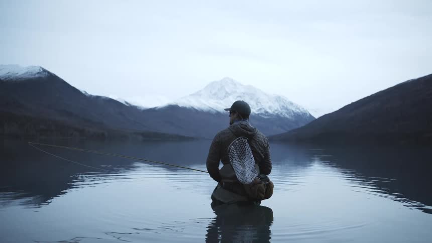 A man wearing a dark jacket and baseball cap kneels in a calm icy lake holding a fishing rod and a net on his back facing a snowcapped mountain peak surrounded by dark slopes