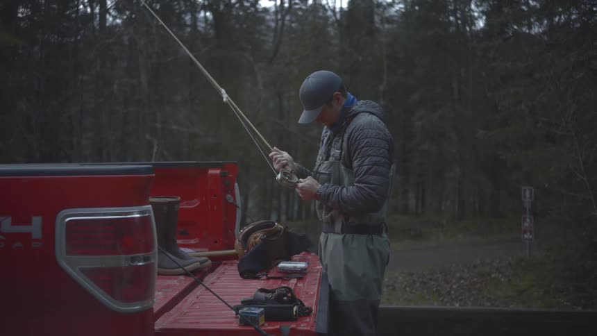 Man prepares a fishing rod on the back of a red pickup truck in a wooded area