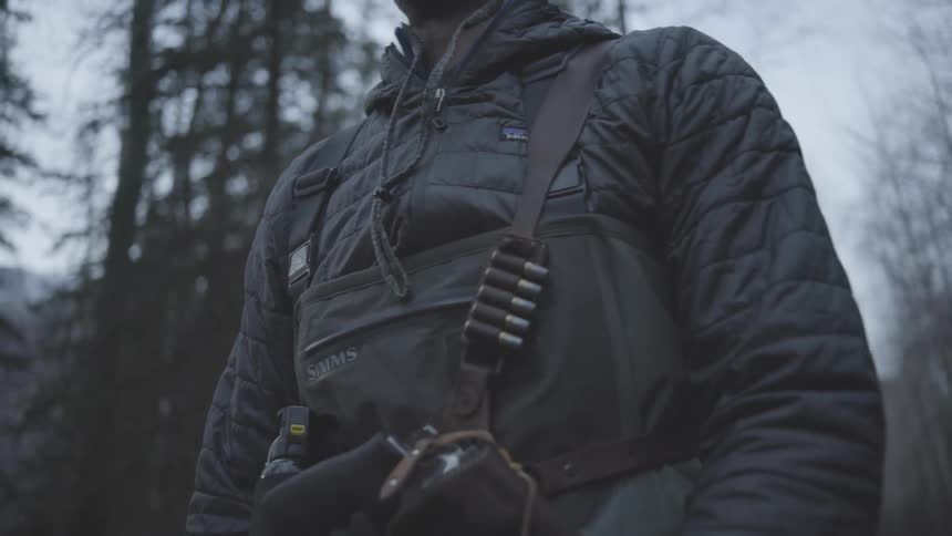 Armed man wearing a cap and jacket walking in a forested area
