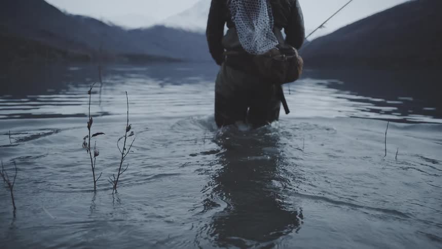 A fly fisherman man wearing dark waders a backpack and carrying a net and rod slowly wades into a cold mountain lake with snowy peaks in the distance