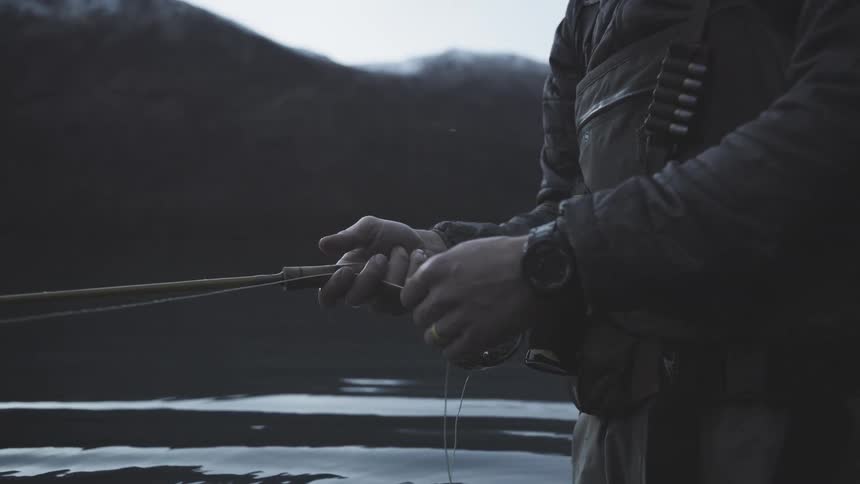 A close view shows a man in dark waders and outdoor gear standing in the dark water of a mountain lake actively holding and reeling a fly fishing rod