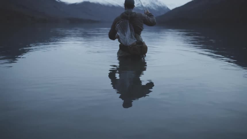A man wades into still water toward snowcapped peaks calm and solitary as he casts his line