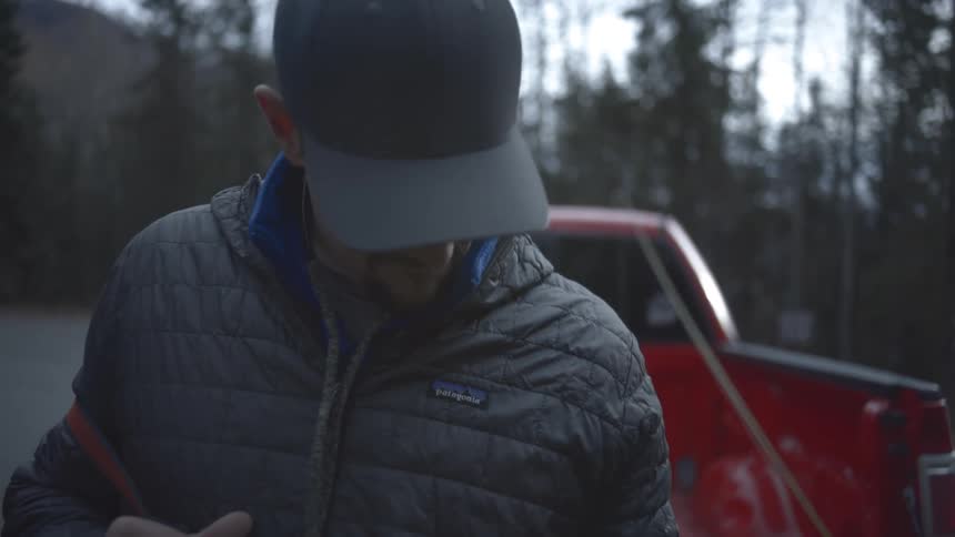 Man adjusts his vest outdoors beside a red pickup truck at dusk
