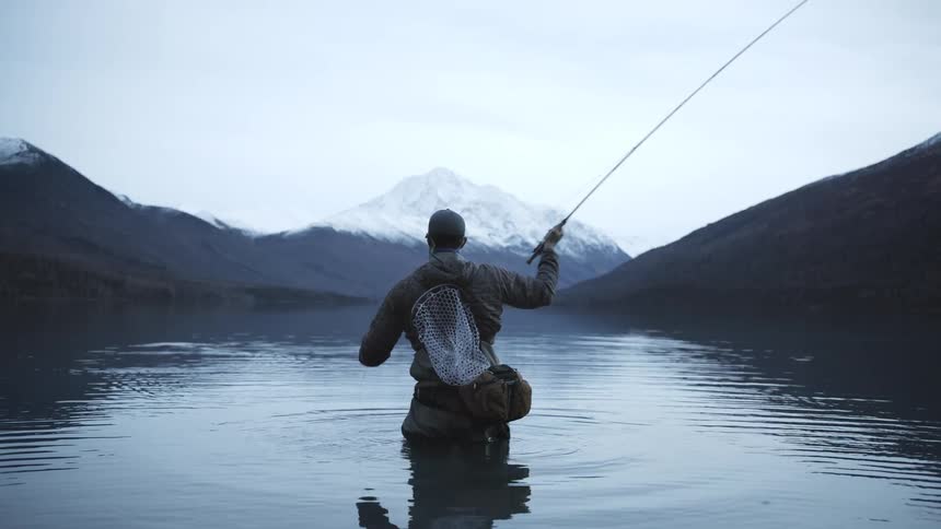 A person wearing waders stands waistdeep in a body of water and repeatedly raises and lowers a fishing rod to cast a fishing line