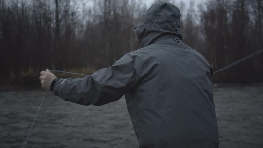 A man in a hooded jacket and tan waders stands in the dark shallow water of a river casting his fishing line toward the dense bare forest on the shore in the dim light