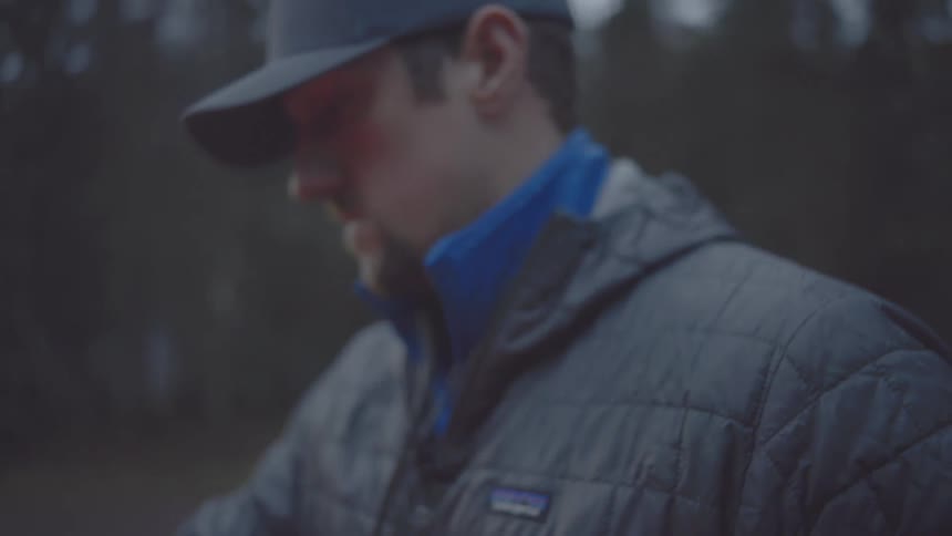 A man in a gray puffer jacket and baseball cap prepares his fishing rod by connecting the two pieces near the open red tailgate of a truck looking out toward a misty lake in the background