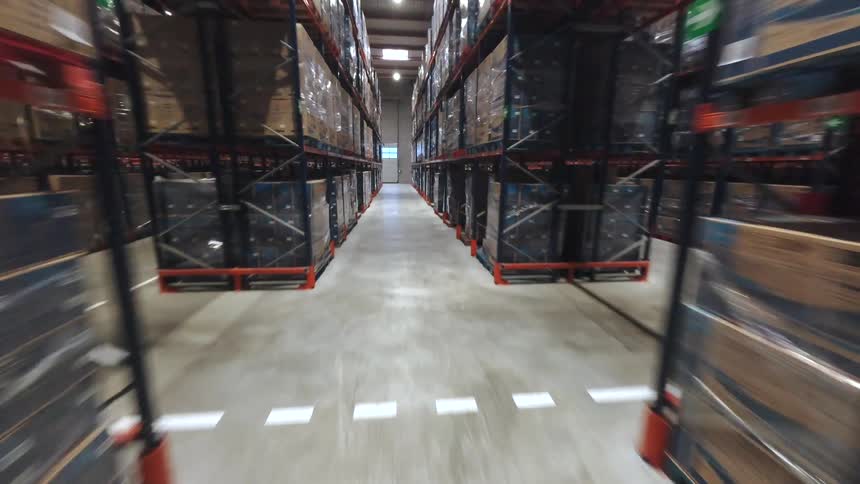 A narrow aisle of a warehouse surrounded by towering shelves stacked with boxed inventory