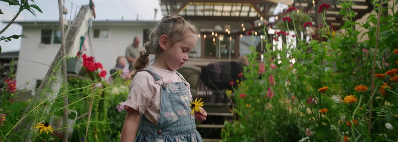 A young girl carrying small garden shears holds a flower that shes picked from one side of a backyard garden then turns around to the other side as on the back porch behind her adults talk