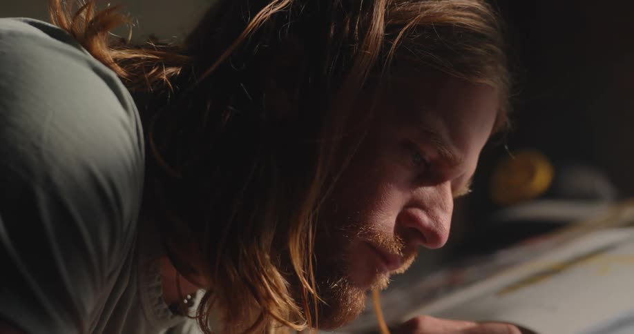 A male artist with long blonde hair and a beard is sketching in a notebook indoors at midday