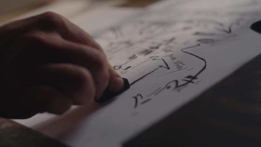 A close lowangle view shows a persons hand holding a dark art tool and actively drawing bold black lines onto a piece of white paper in a dimly lit setting