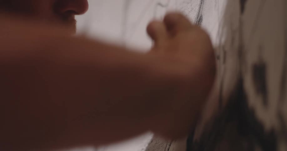 A close view shows a mans hand with a dark art tool actively drawing bold black curves onto a large white canvas in a welllit studio