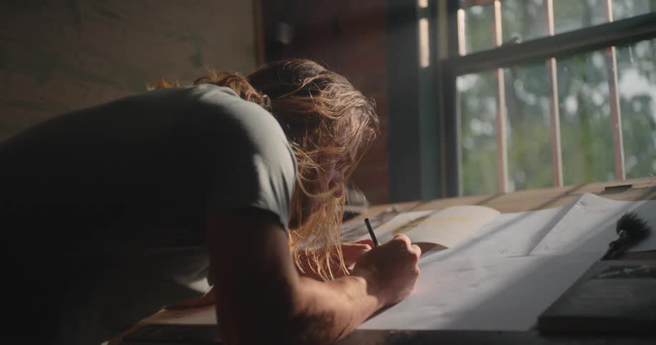 A young man leans over a large desk holding a pencil and write on a paper