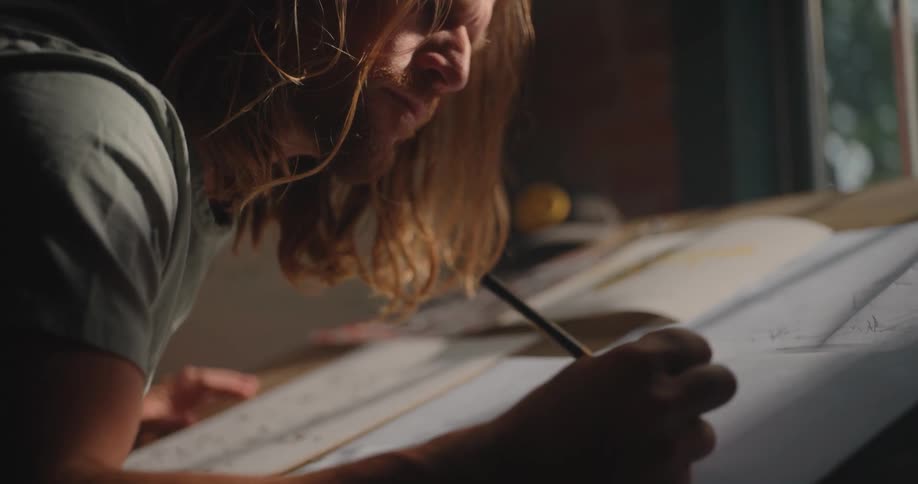 A male artist with long blonde hair and a beard is sketching in a notebook with a pen indoors at midday