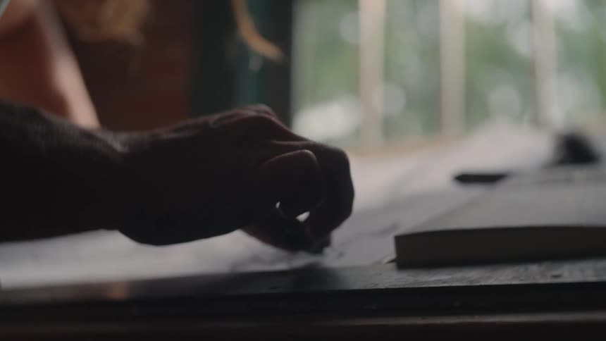 A darkly silhouetted hand of a person sketches with a pencil on a sheet of white paper on a table with a bright window and outdoor greenery visible in the blurred background