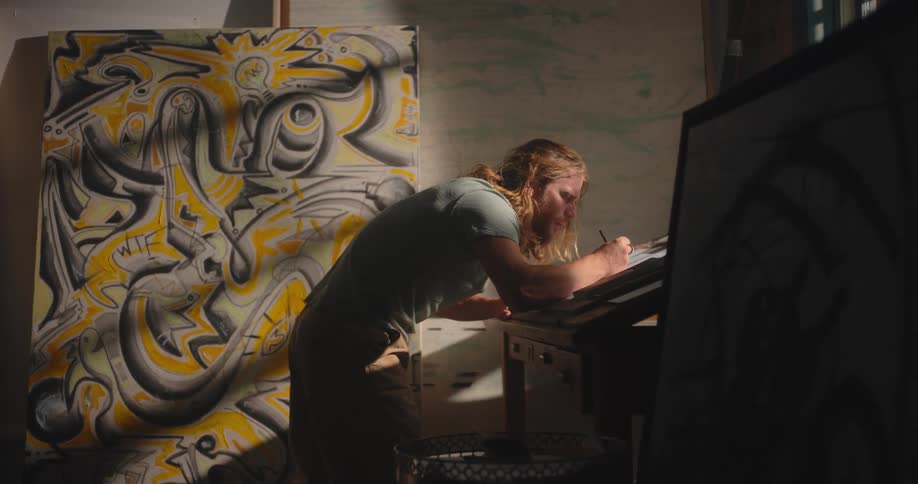 An artist with long hair and a beard sketches intently at a drafting table in a dimly lit studio with a large abstract yellow and black painting in the background