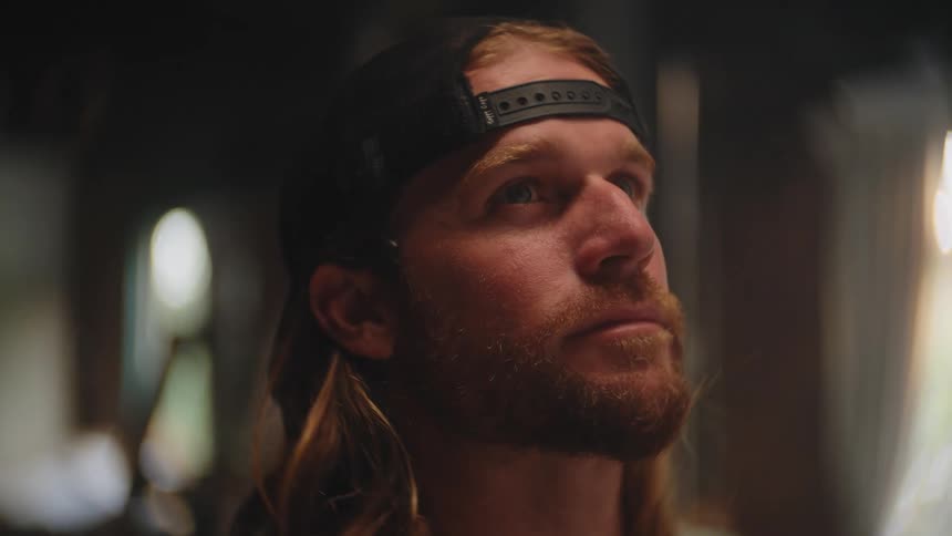 A close view shows a redbearded man with long blonde hair and a black backward baseball cap looking up intently in a dark interior space with bright background highlights