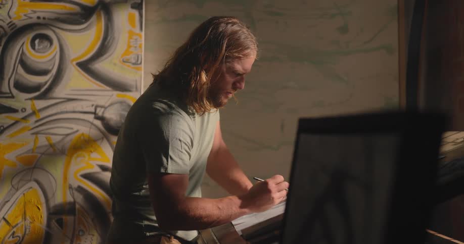 Man drawing on a tilted desk in an art studio with paintings in the background