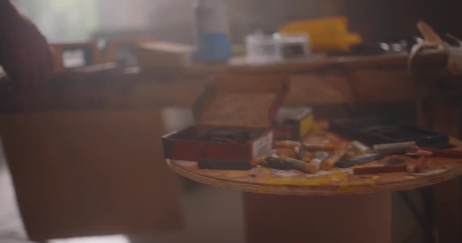 A persons hands moving a yellow and black level tool and other objects around a table that is cluttered with various art tools and materials