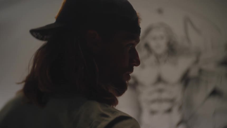 He video is a closeup profile shot of an artist looking intently at a large wall drawing behind himthe drawing which is the blurred background is a charcoal depiction of a muscular figure resembling the vitruvian man