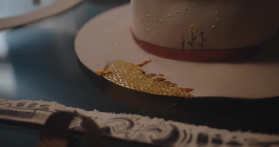 The video is an extreme closeup focusing on the brim of a lightcolored hatthe hat is being decorated with a small goldenyellow decorative piece possibly a dried leaf or resin element near an embroidered flower design