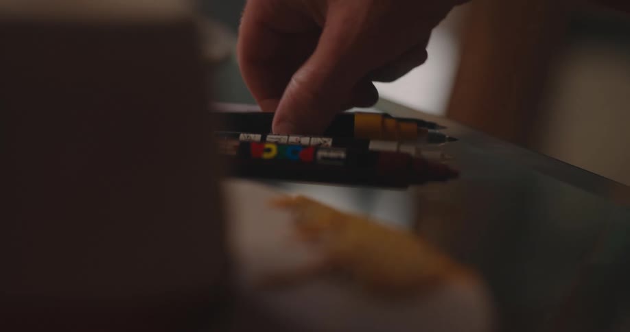 A hand reaches in and subtly adjusts a small lowlying pile of marker pens resting on a dark reflective surface which may be a glass table