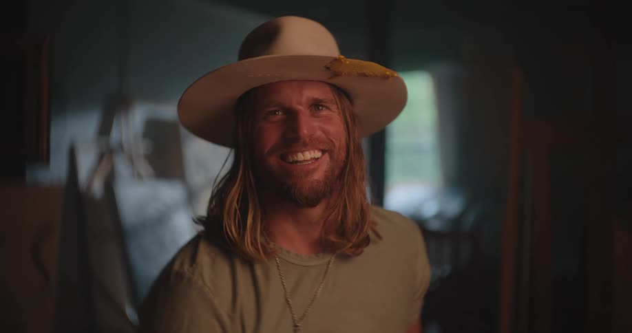 The video is a medium closeup of a smiling man with long blonde hair and a beardhe is wearing a lightcolored fedorastyle hat with a decorative feature and an olive green shirt with a pendant necklace