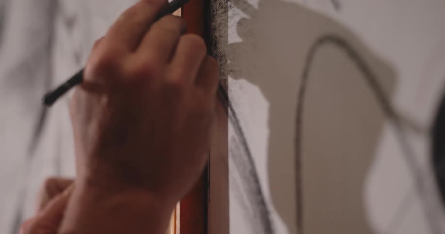 The video is an extreme closeup of a persons hands working on a large drawing or muralthe artist is using a pencil or piece of charcoal along the edge of a wooden straightedge or ruler to create a bold straight line on the white paper or