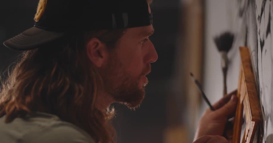 A young man with a beard and long hair wearing a cap meticulously holding a wooden triangle against a surface and using a dark pencil to draw in the wall