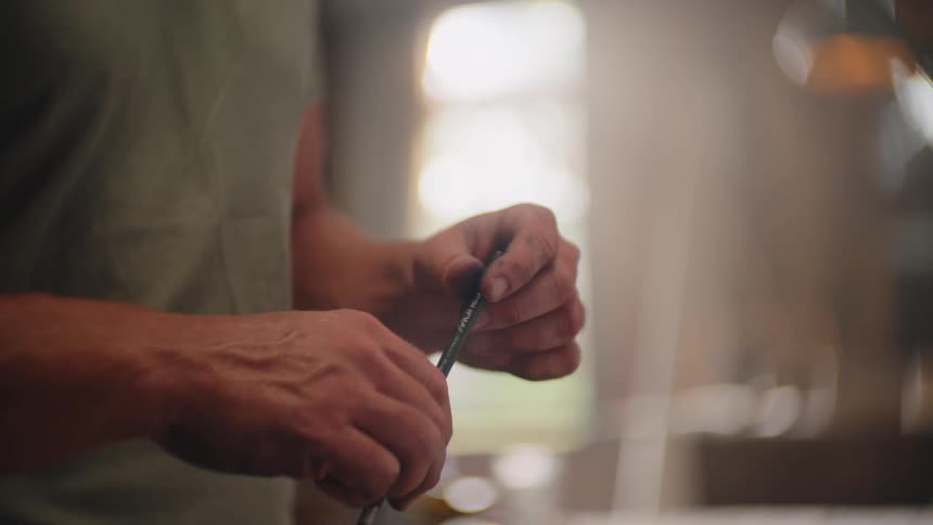 A close view shows the hands of a man wearing a green shirt twisting a pencil in a brightly lit indoor workspace