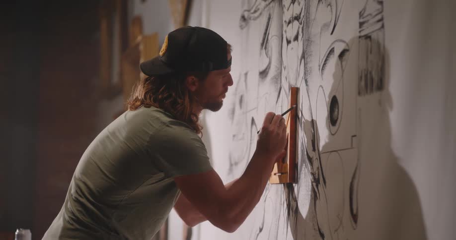Close up of a male artist finishing a large drawing on a white canvas on the wall in a room during daytime