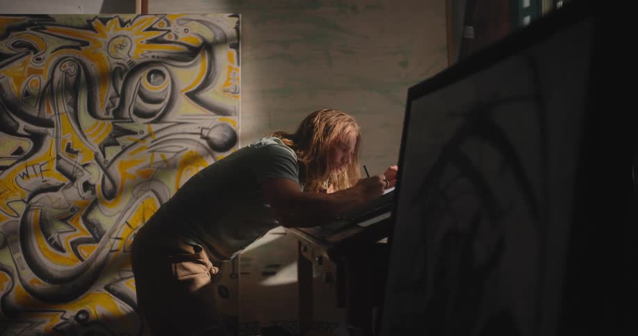 A man is leaning over drawing on paper surrounded by large paintings in an art studio