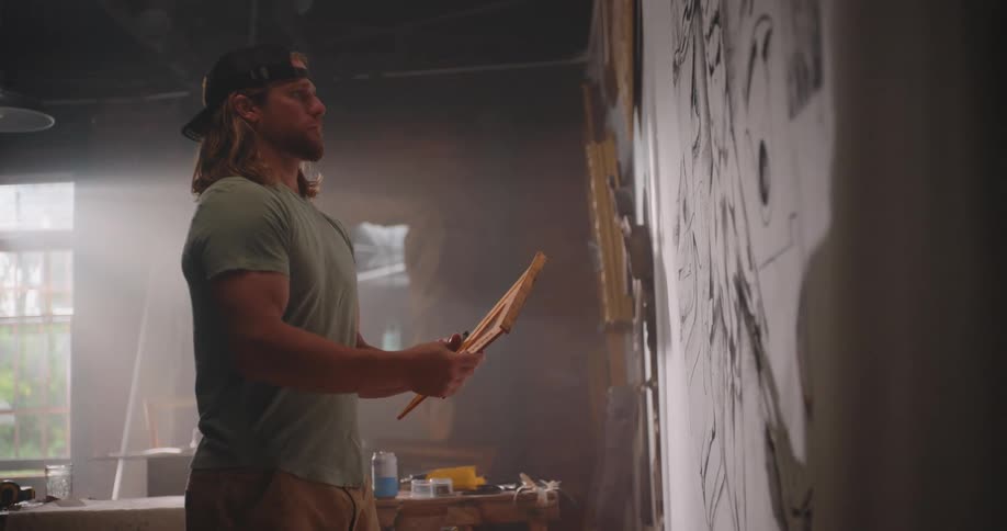 A man with long hair and a baseball cap standing in a studio picking up a ruler and a set square from a workbench then holding the tools against a large sheet of paper on the wall to measure a drawing