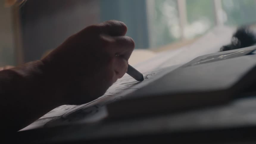A close angled view shows a hand holding a dark art crayon or charcoal stick sketching on white paper at an angle near a bright window