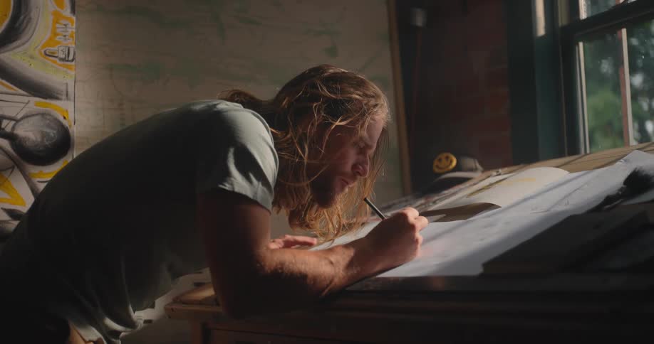 A man with long hair leaning over a slanted wooden desk near a window using a tool to draw on paper