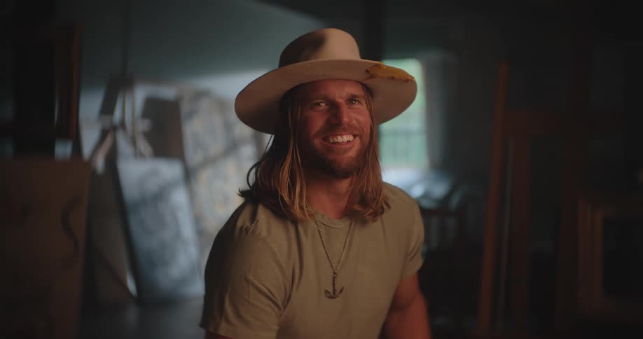 A man sitting in a studio with a beard and long hair wearing a hat his shoulders moving as he laughs surrounded by canvases and paintings leaning against the walls