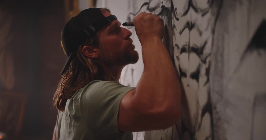A close view shows a muscular man with long brown hair and a black backward baseball cap drawing intently with a dark art tool on a large black and white mural on the wall
