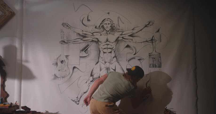 A wide shot shows an artist wearing a cap and lightcolored clothing drawing on a large piece of paper or canvas mounted on a wallthe drawing is a large stylized rendition of the vitruvian man figure surrounded by abstract sketchy lines and shapes