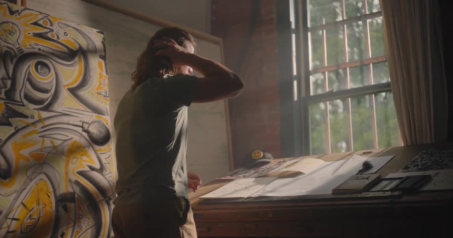 A longhaired man in a green shirt and tan pants leans over a drafting table drawing on paper next to a large yellow and black canvas as bright daylight streams in through a tall window