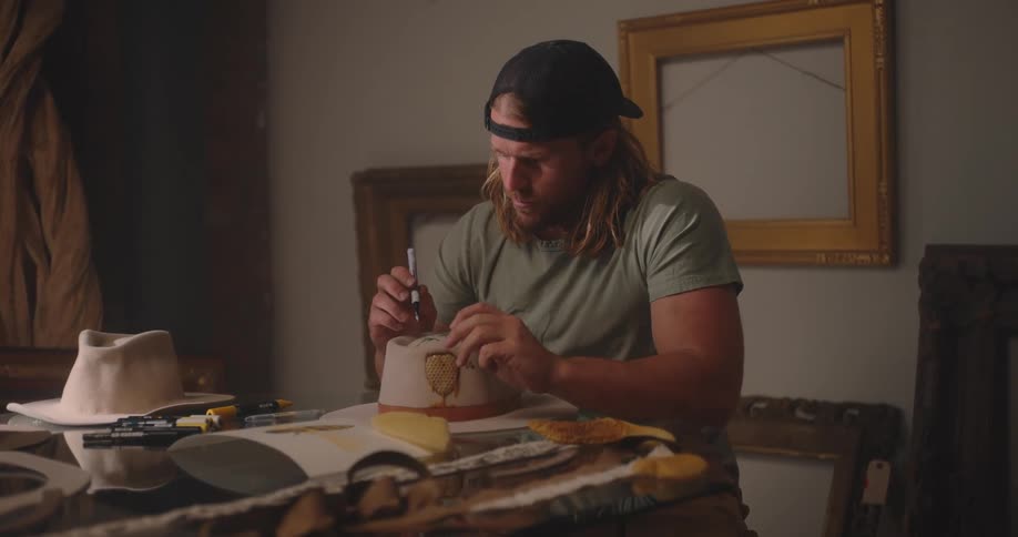 Man wearing a cap works on customizing a hat in a studio surrounded by frames