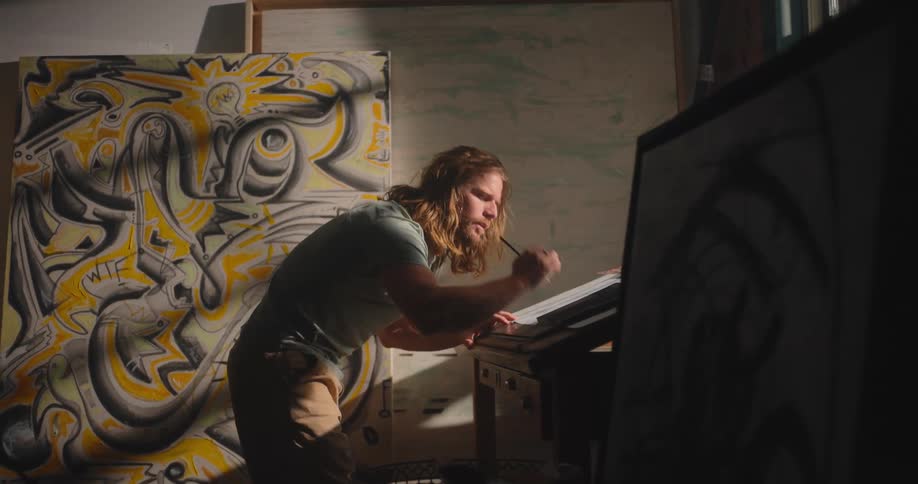 Man draws with focus in a studio surrounded by large colorful canvases