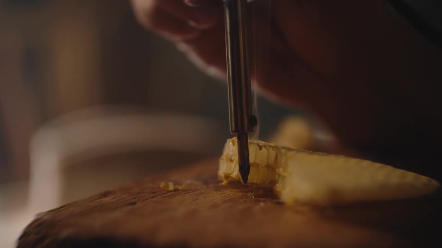 A close view shows the hand of a person using a heated metal tool to melt and remove the capping wax from a piece of golden yellow honeycomb resting on a wooden surface