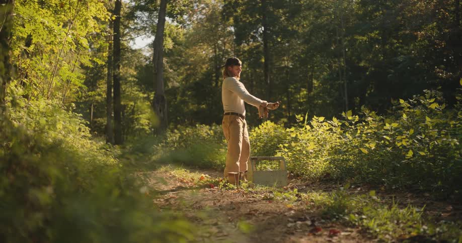 The video is a wide shot of a man standing in a sundappled overgrown path surrounded by forest greeneryhe is wearing a lightcolored longsleeve shirt and tan pants and he is putting on a pair of gloves over what appears to be a metal contai