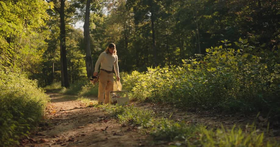 Beekeeper walking along a wooded path carrying equipment under soft light