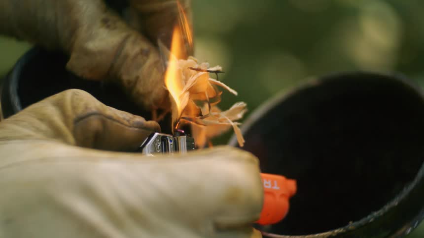 The video is a closeup shot of gloved hands using a lighter to ignite wood shavings in preparation for a firethe focus is on the small flame catching the tinder as it is held over a dark cylindrical container likely a beekeepers smoker