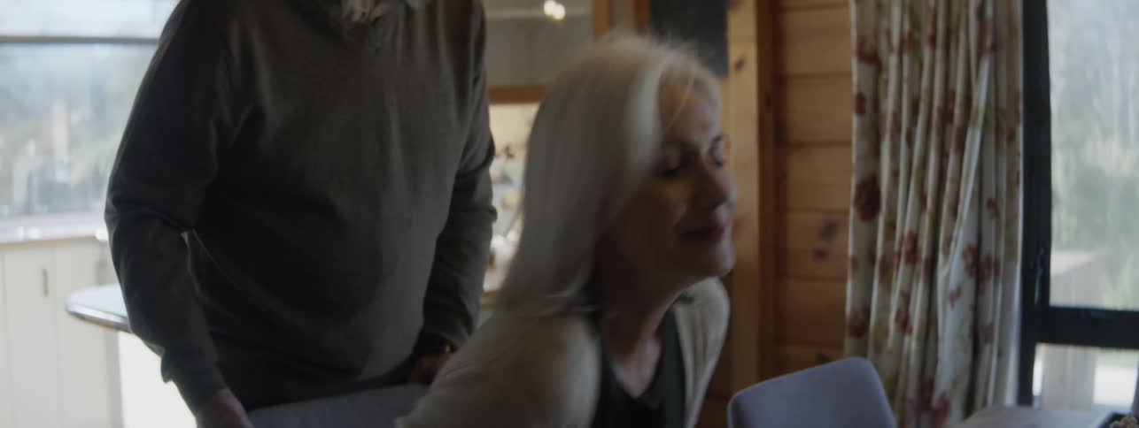 An older woman with white hair wearing a tan cardigan smiles while interacting with an older man with a grey beard in a bright wooden kitchen with large windows
