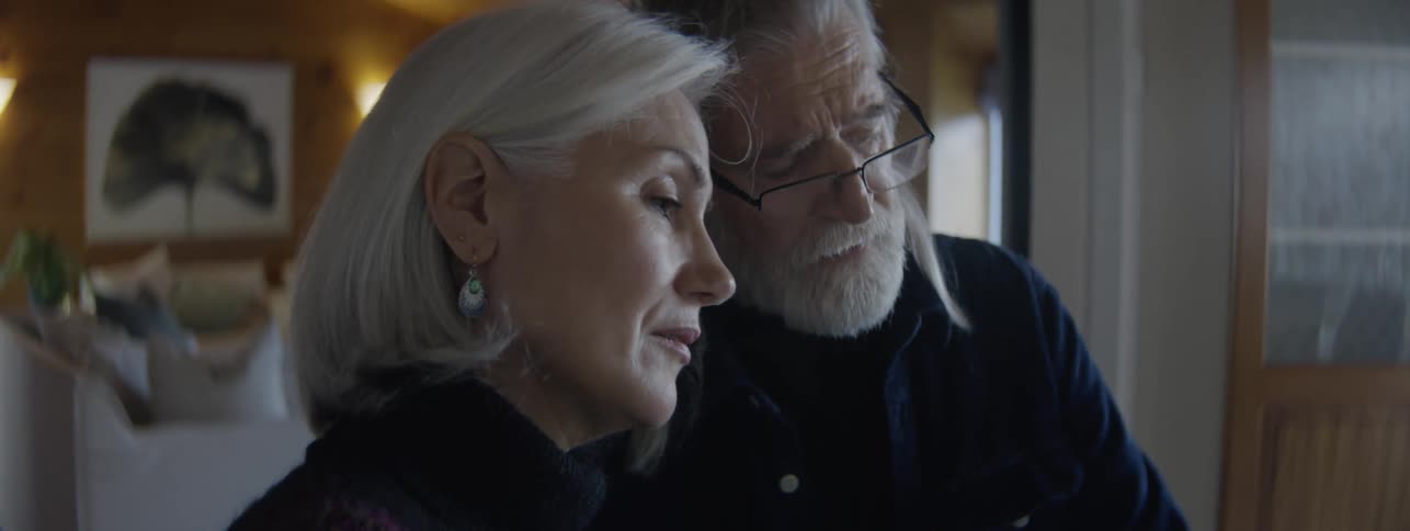 A close view shows an older man with a white beard and glasses gently leaning his head on the head of an older woman with white hair and earrings for a loving moment in a softly lit room