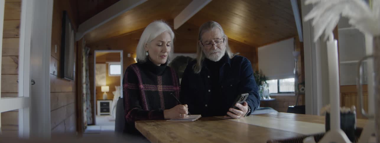 An older man with long white hair and glasses holds a smartphone as an older woman with white hair writes in a notebook at a wooden table in a cozy woodpaneled room