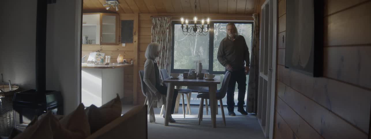 An elderly couple sets food on the dining table to have breakfast together then both sit down afterward to eat