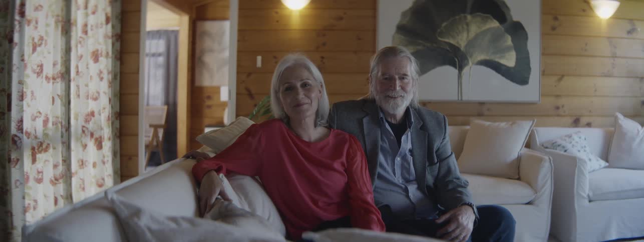 The video shows an elderly couple sitting together on a white sofa in a welllit living room with woodpaneled wallsthey are both looking directly at the camera with a gentle smile the woman is wearing a red top and the man is wearing a blaze