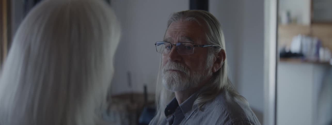 A closeup camera shot shows the profile of an older man with long gray hair a beard and glasses looking at a person whose back of the head is in the foreground
